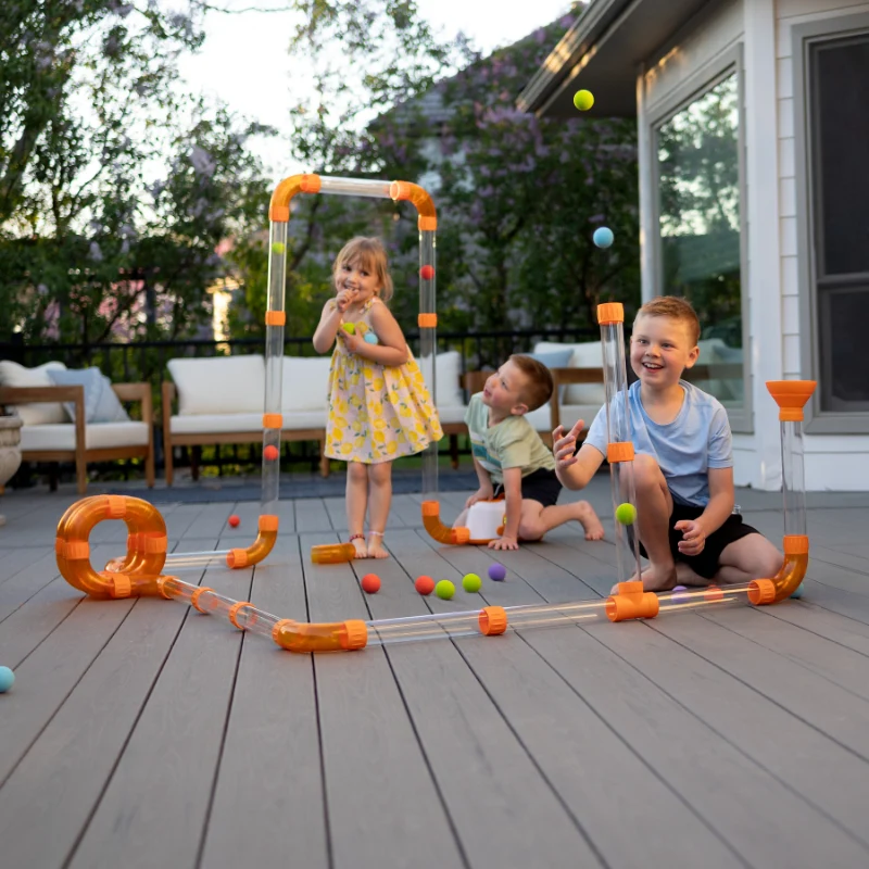 Drie kinderen spelen op een terras met doorzichtige luchtbuizen, oranje bochten en gekleurde schuimballen